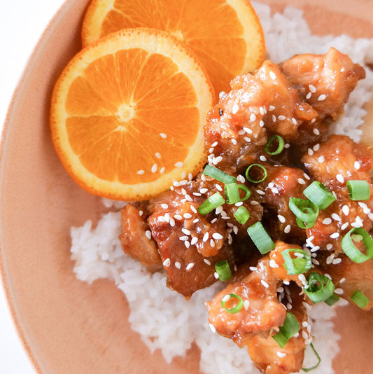 Bowl of orange chicken served over rice, topped with sesame seeds, green onions, and orange slices