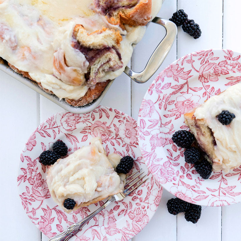 A serving of blackberry cinnamon rolls topped with frosting, served on floral plates with fresh blackberries.