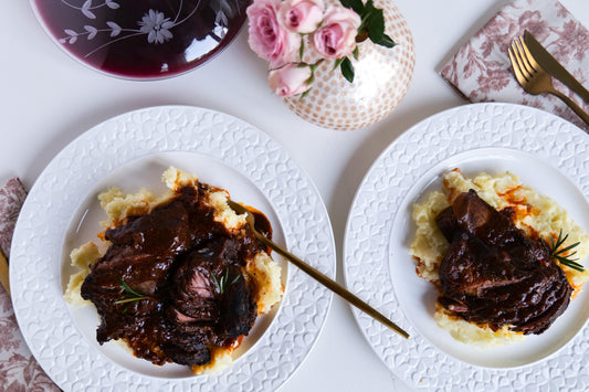 Two plates of braised short ribs served over mashed potatoes with a side of pink roses and a glass of red wine.