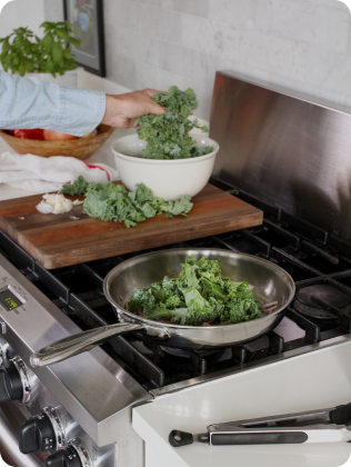 A person adding kale to a pan on a stove, with a bowl of kale and garlic nearby.