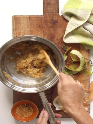 A hand stirring cooked onions in a pot on a cutting board with spices and peels nearby.