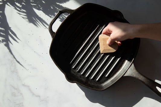 A hand holding a natural wood pan scraper over a cast iron grill pan