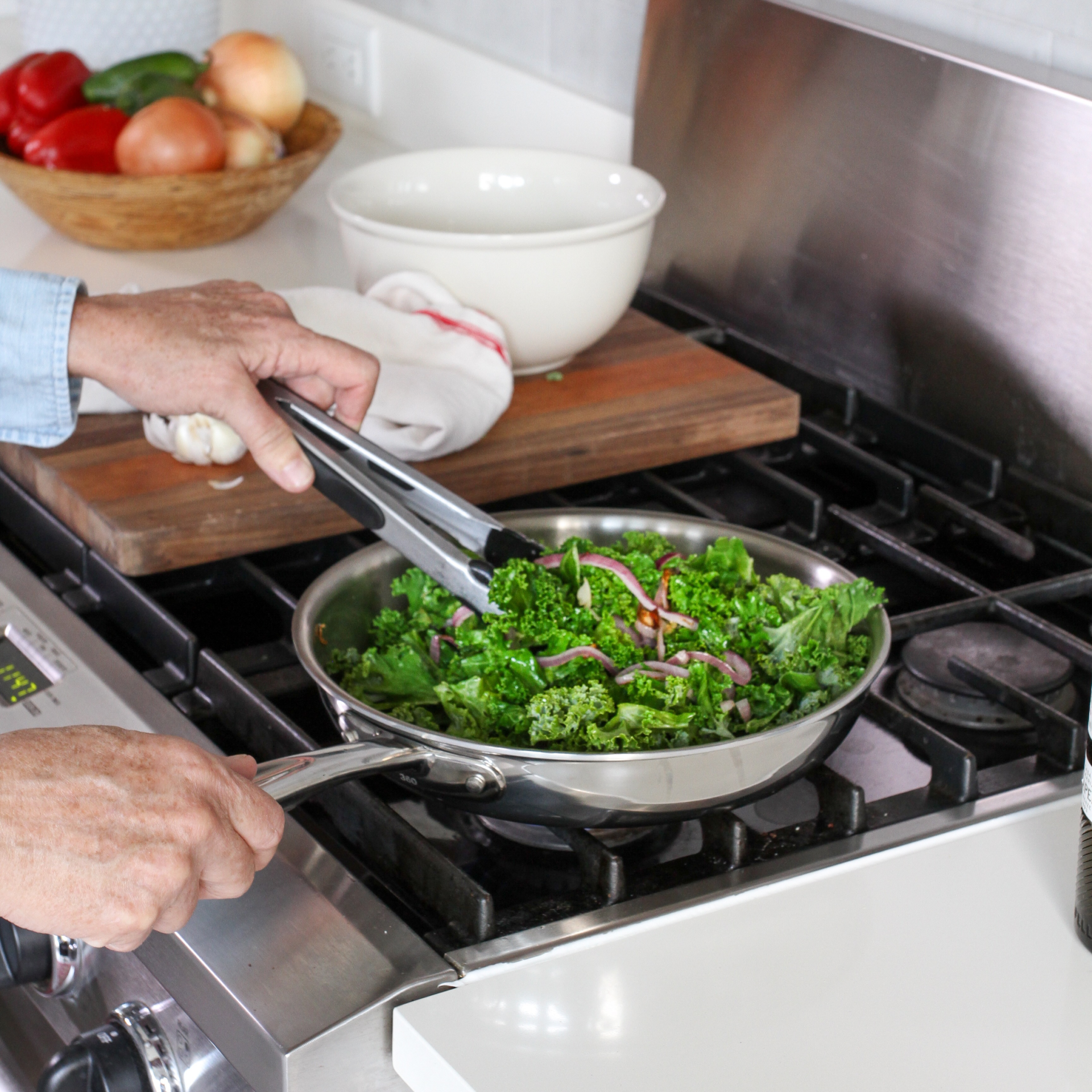 A person using tongs to stir kale and onions in a stainless steel fry pan on a stove.