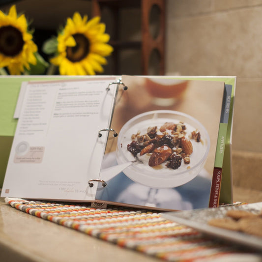Open cookbook page showing a dish with nuts and dried fruits on a table with sunflowers in the background.