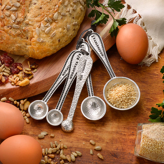Set of five pewter measuring spoons with engraved measurements, surrounded by eggs, seeds, and bread.