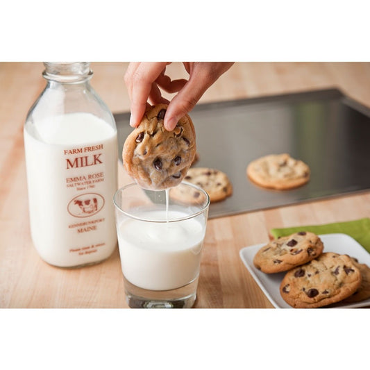 A hand dipping a chocolate chip cookie into a glass of milk, with cookies on a tray in the background.