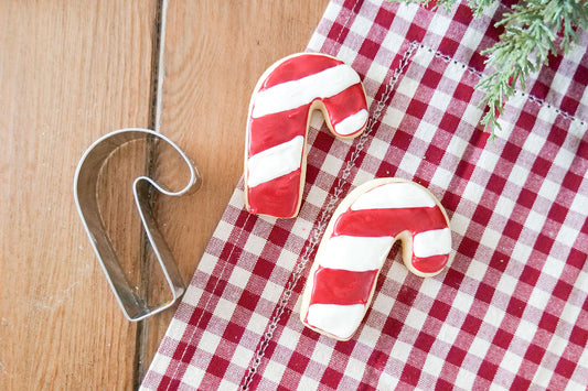 A 3 1/2" candy cane cookie cutter next to decorated candy cane cookies on a checkered tablecloth.