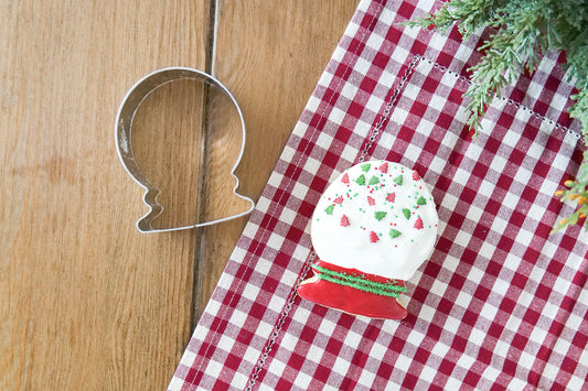 Snowglobe cookie next to a snowglobe-shaped cookie cutter on a checkered tablecloth