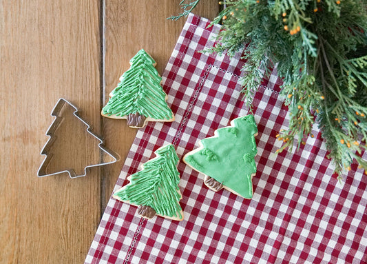 Three decorated Christmas tree cookies next to a Christmas tree cookie cutter on a checkered tablecloth.