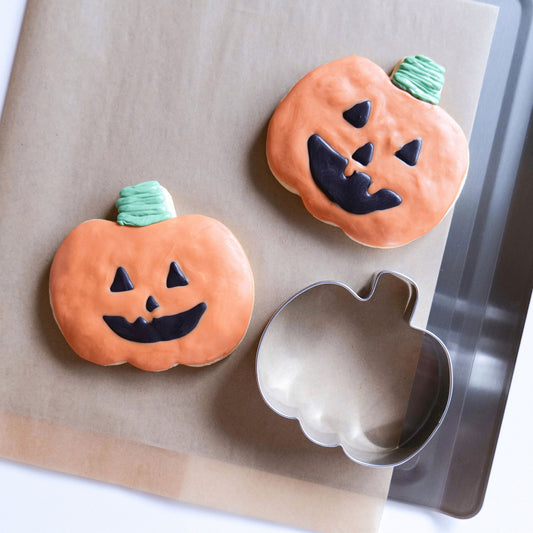 Two decorated pumpkin cookies and a pumpkin-shaped cookie cutter on a baking sheet.