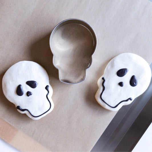 Two decorated skull cookies with a skull cookie cutter on parchment paper.