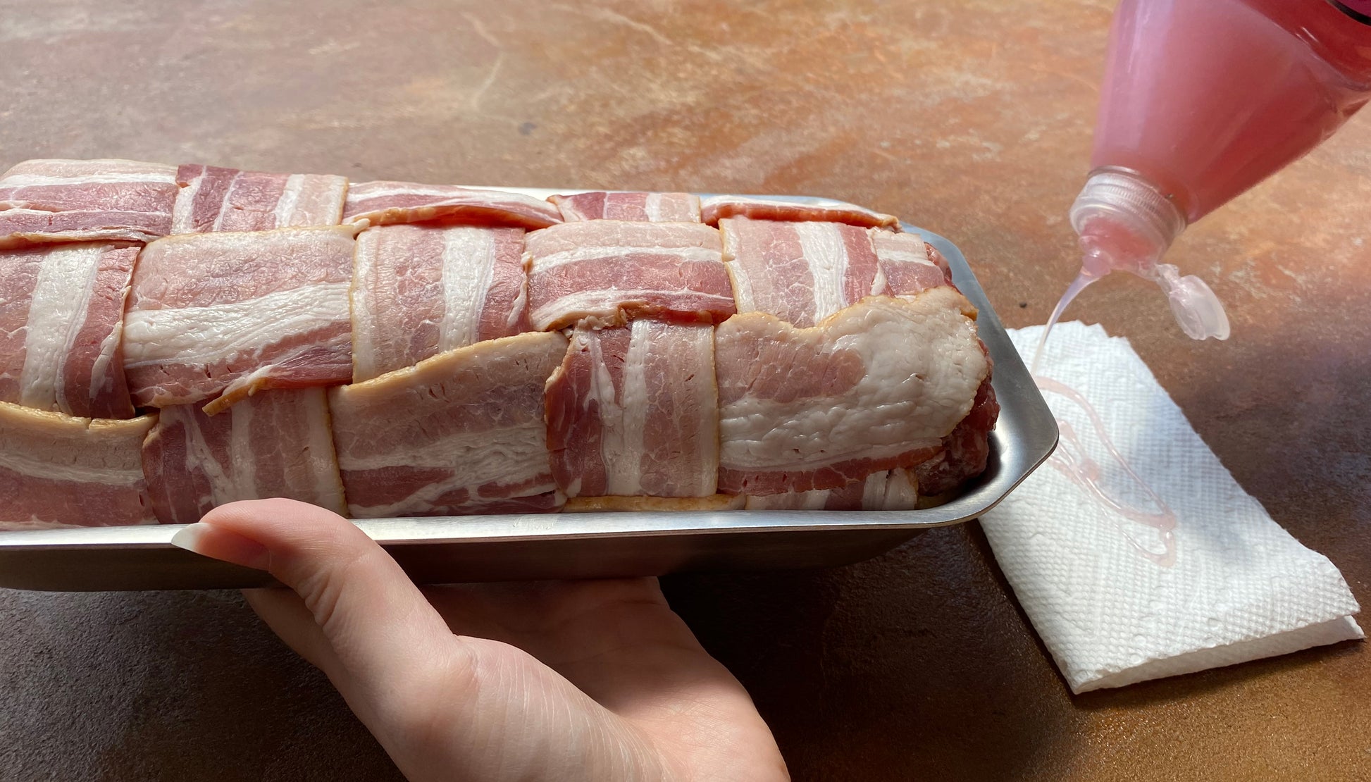 A hand holding a mini jelly roll pan filled with bacon-wrapped meatloaf, with a bottle pouring sauce nearby.