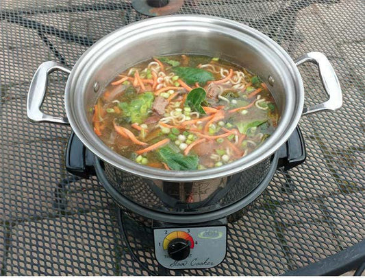 Top-down view of stainless steel 4-quart stockpot filled with beef Asian noodle soup on a slow cooker base on a metal mesh patio table