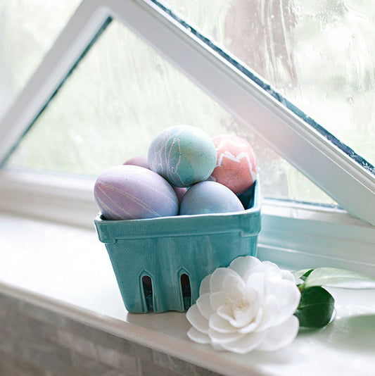 Pastel-dyed Easter eggs arranged in a turquoise berry basket on a windowsill next to a white camellia