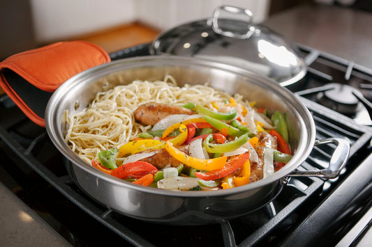 Stainless steel pan on stove with cooked noodles, sausages, and sliced red, yellow, and green bell peppers