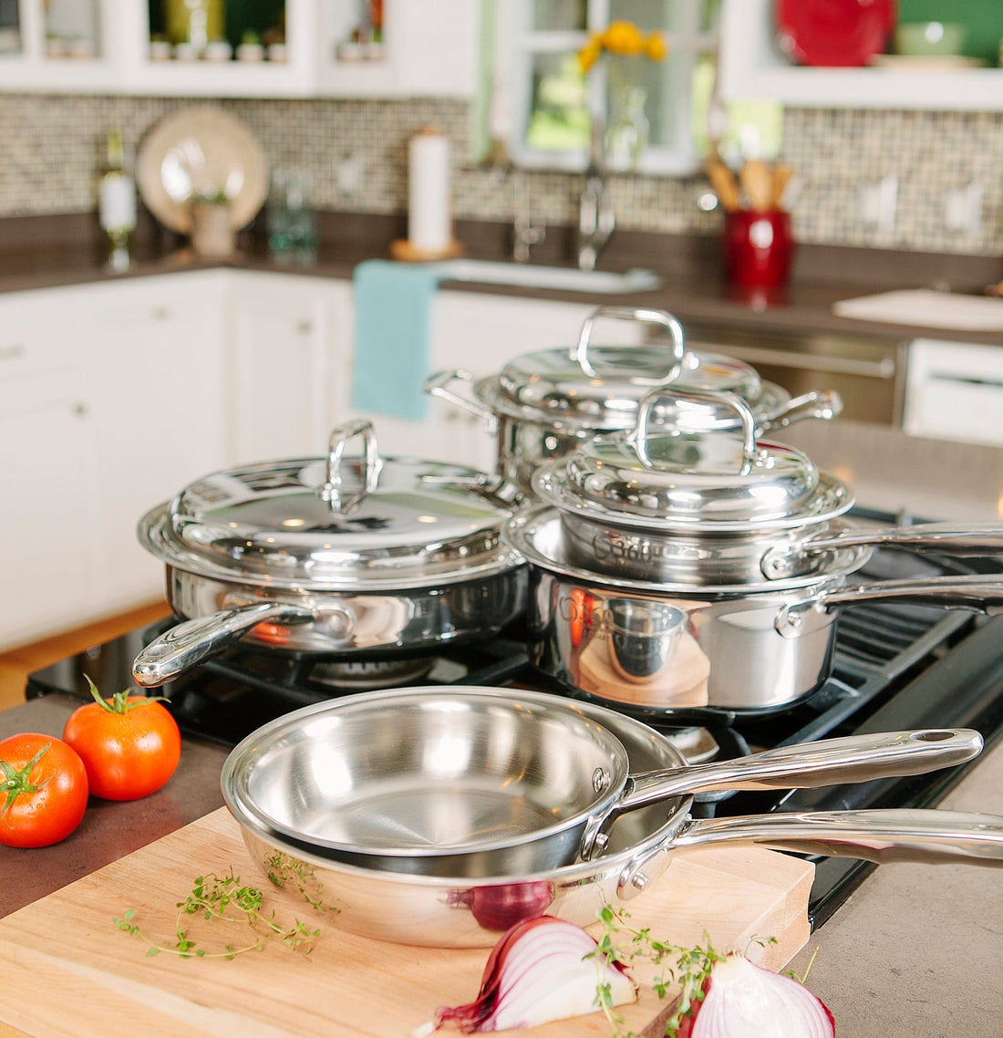 Polished stainless-steel pots and pans on a gas stove, with tomatoes, sliced red onion, and herbs on a cutting board.