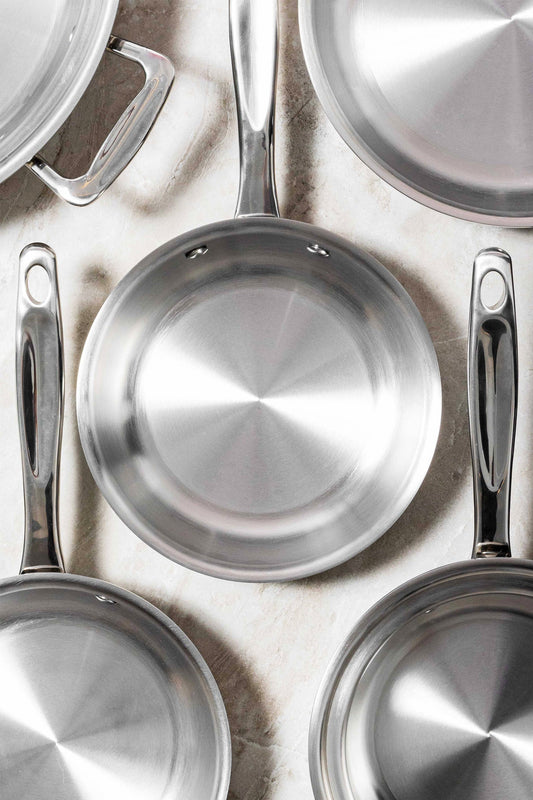 Top-down view of stainless steel frying pans showcasing polished interiors and riveted handles on a light stone surface