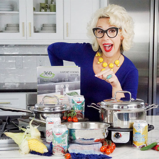Chef smiling behind 360 Cookware pots and pans on a kitchen counter with corn, cherry tomatoes, canned drinks, and a sign reading Vapor Cooks Better