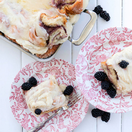 A serving of blackberry cinnamon rolls topped with frosting, served on floral plates with fresh blackberries.