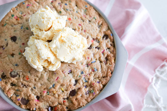 Top-down view of pizookie in a round skillet topped with three scoops of vanilla ice cream, studded with chocolate chips and rainbow sprinkles on a pink cloth