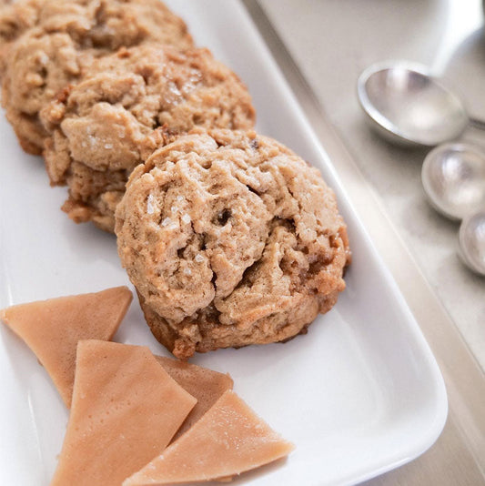 Three brown sugar toffee cookies on a white rectangular platter with broken toffee pieces and metal measuring spoons in the background