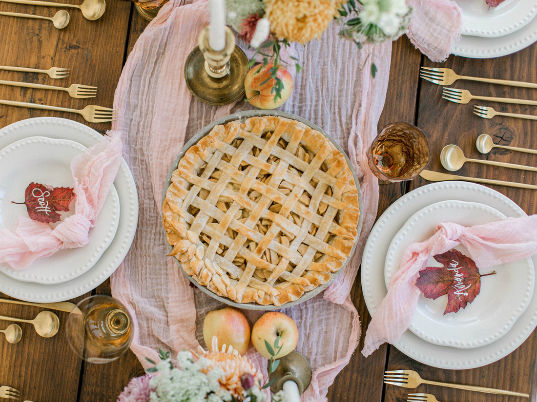 Lattice-top apple pie on a pink gauze runner, surrounded by white place settings, gold flatware and apples