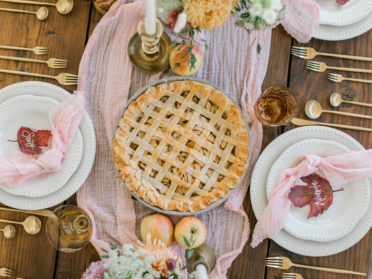 Lattice-crust apple pie on a wooden table with pink cloth runner, surrounded by white plates with pink napkins and autumn leaves labeled 'Grat' and 'Thankful', gold cutlery, and apples.