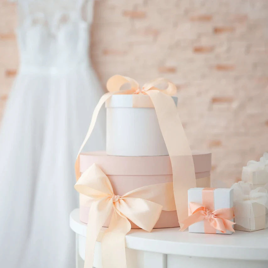 Stacked round and square gift boxes with peach satin bows on a white table; blurred wedding dress in background