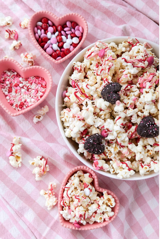 Bowl of white chocolate popcorn drizzled with pink and red chocolate, surrounded by heart-shaped bowls of candy and sprinkles.
