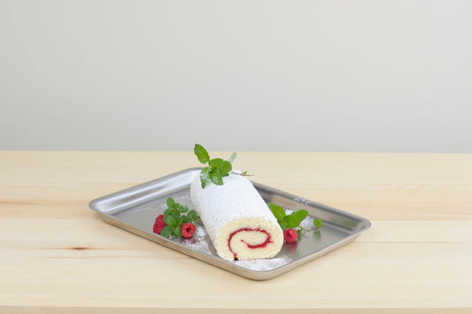 Angled view of a stainless-steel jelly roll pan holding a powdered-sugar raspberry jelly roll garnished with fresh raspberries and mint on a light wood table