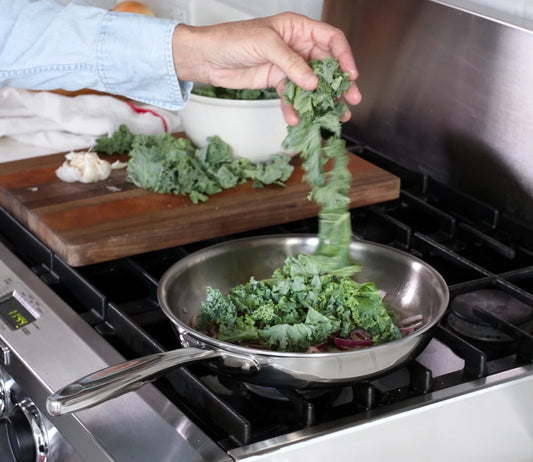 A hand adding kale to a stainless steel frying pan on a stove with chopped garlic and a bowl of kale in the background.