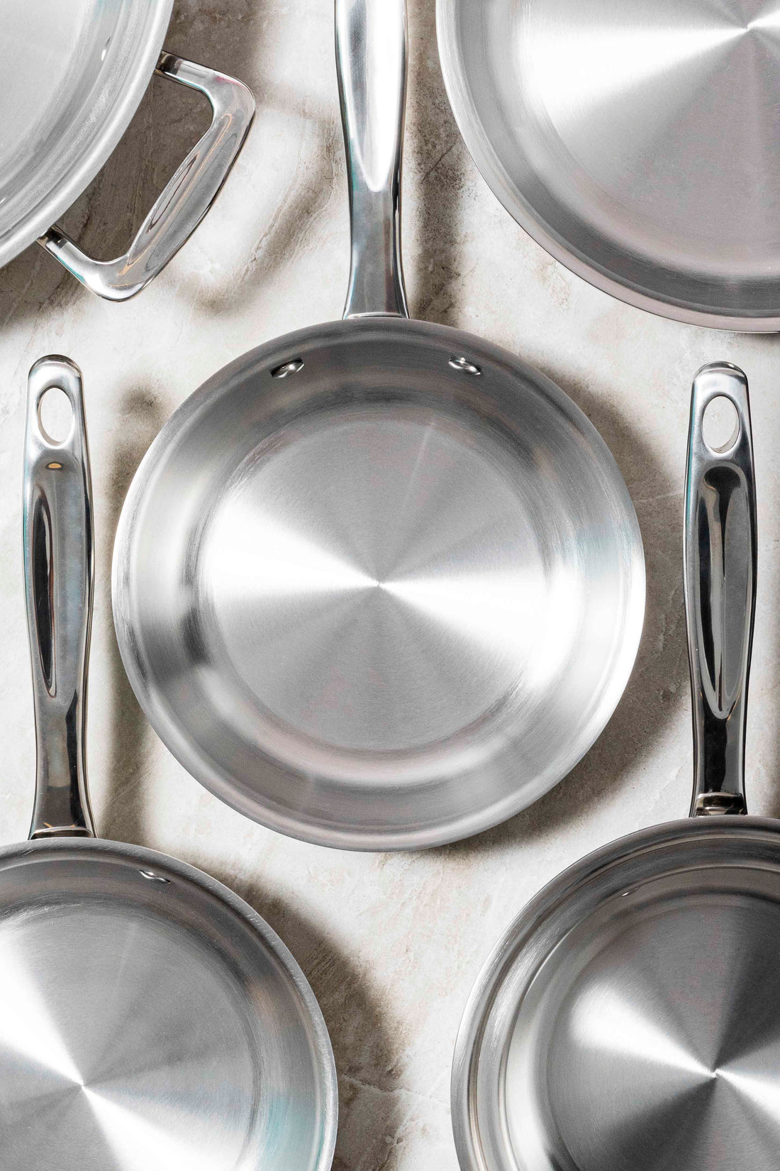 Top view of stainless steel frying pans showing polished interiors and fitted handles on a light marble background
