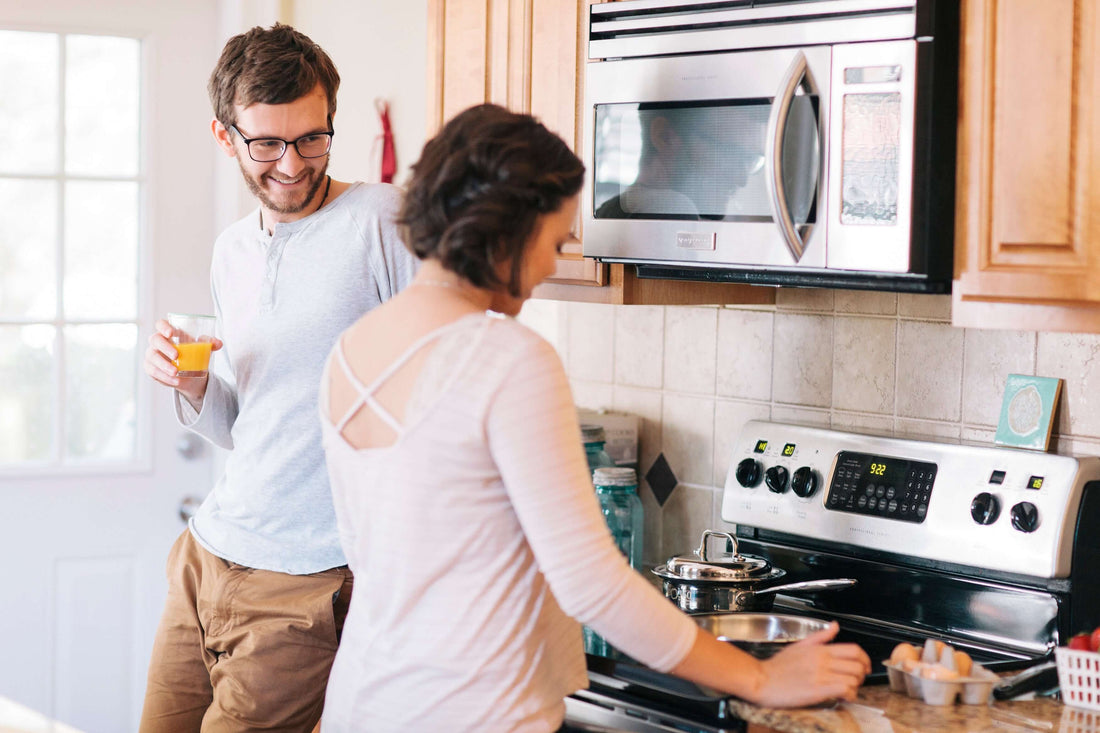 Man holding a glass of orange juice smiling at a woman cooking on a stove in a bright kitchen with wooden cabinets and stainless appliances