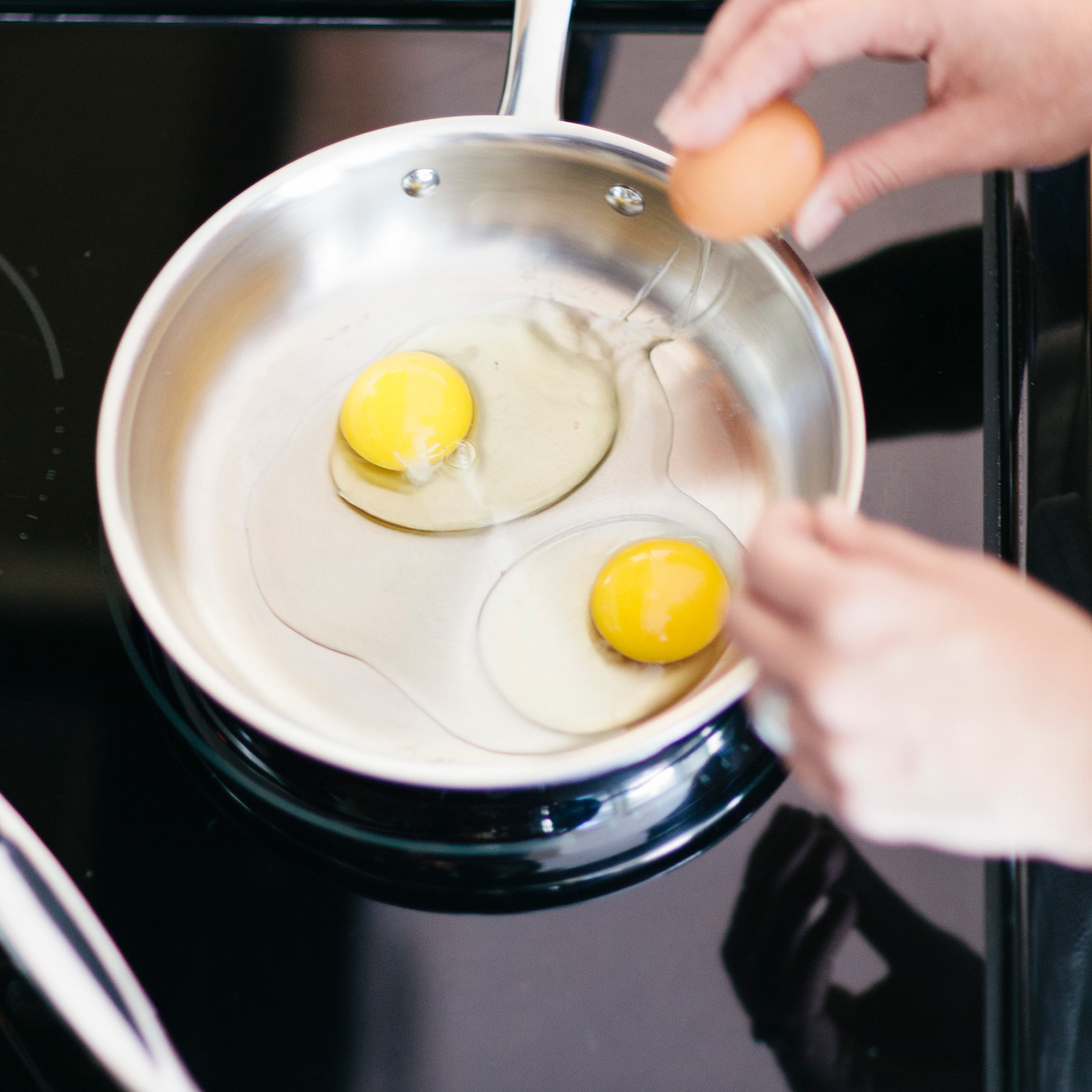 Two eggs being cracked into a stainless-steel frying pan on a stovetop, hands holding an egg and a fork.