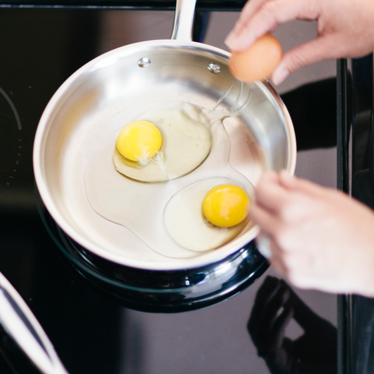 Two eggs being cracked into a stainless-steel frying pan on a stovetop, hands holding an egg and a fork.