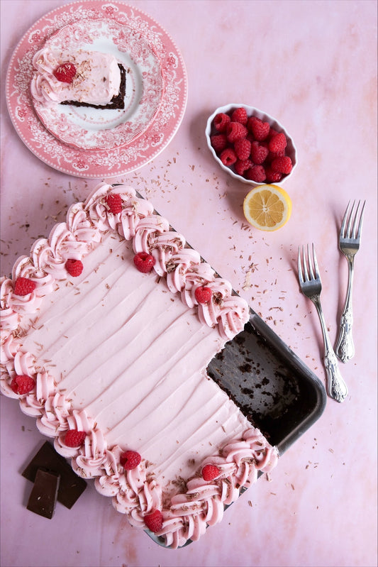 Espresso chocolate cake topped with raspberry cream cheese frosting, garnished with raspberries and chocolate shavings, served with a slice on a plate.