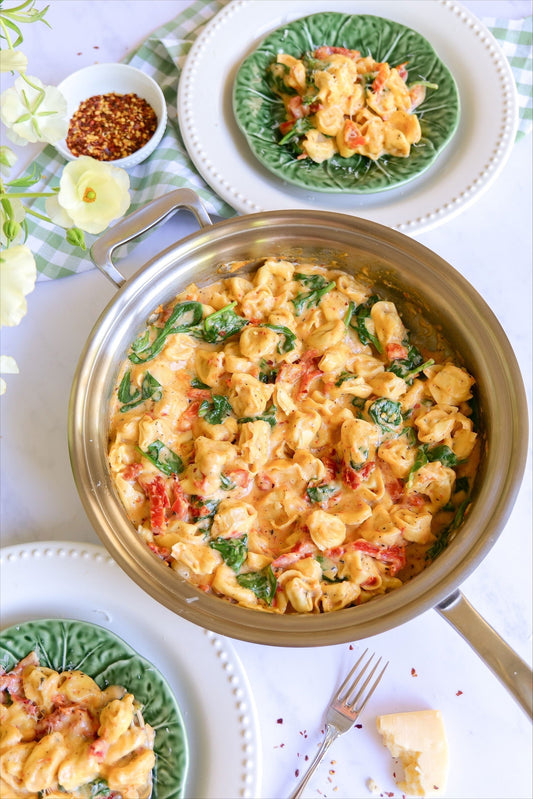 Creamy tortellini with spinach and sun-dried tomatoes in a stainless-steel pot on a white table setting