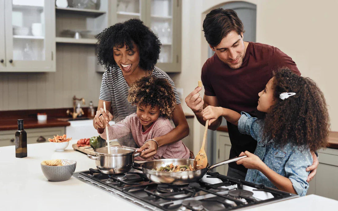 Parents helping two children cook on a kitchen island, stirring food in stainless-steel pans on a gas stove