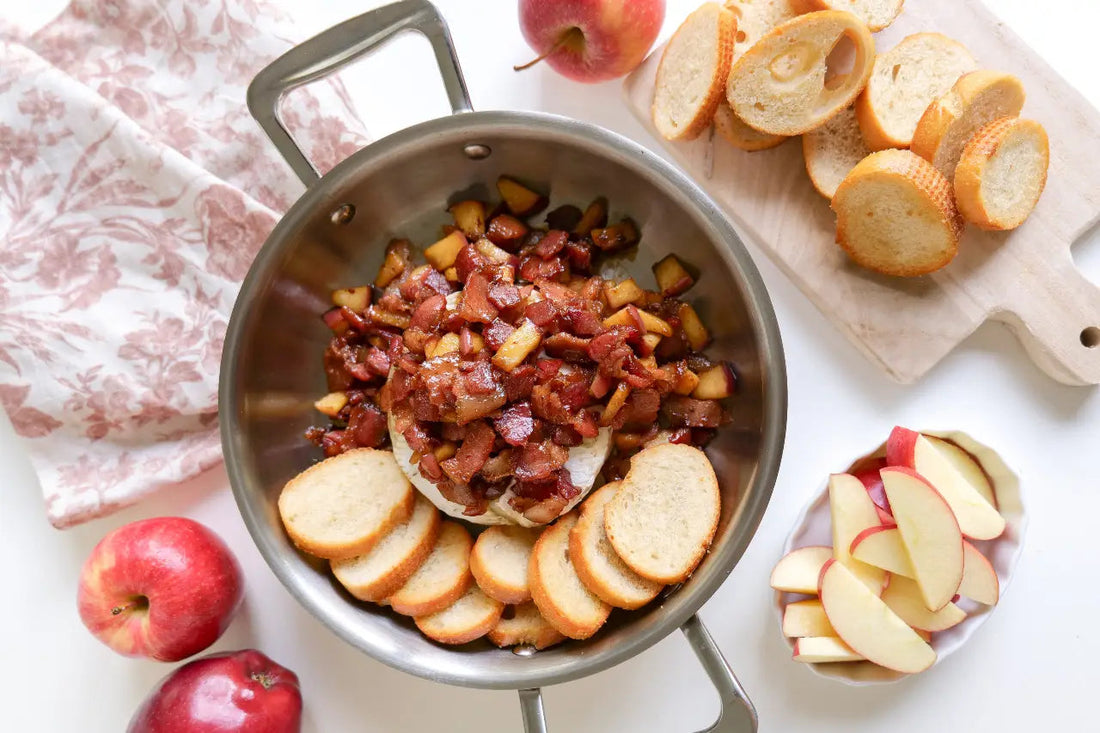 Baked brie in a pan topped with bacon and diced apples, surrounded by sliced baguette rounds and apple wedges.