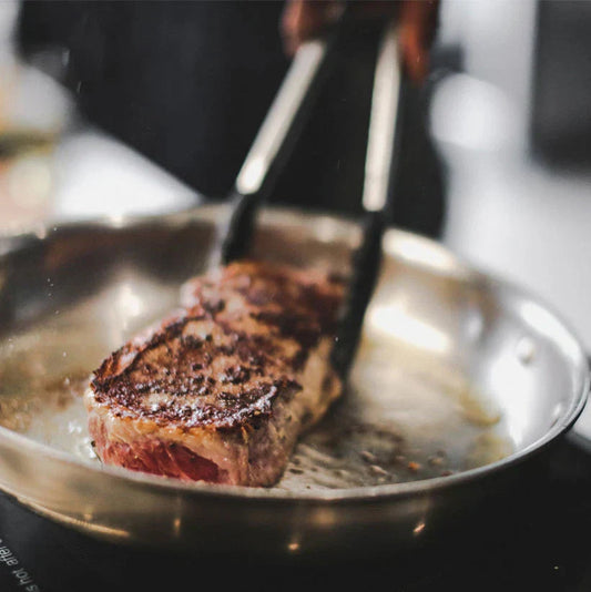 Tongs lifting a seared steak in a stainless steel pan with browned fond and cooking juices