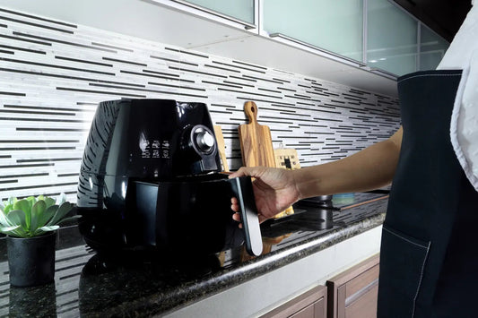 Person pulling open a black air fryer basket on a kitchen counter with cutting boards and a small potted succulent in the background