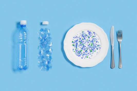 Plate of blue-and-green microplastic flakes beside a shredded plastic bottle and knife and fork on a blue background, illustrating microplastic contamination of food