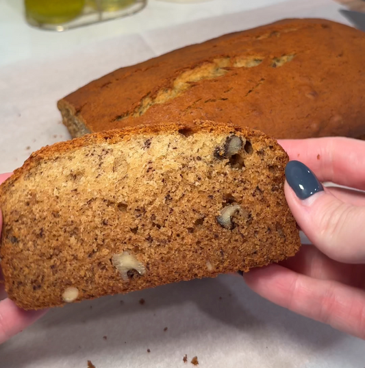 Close-up of a slice of walnut banana bread held by hands, showing moist crumb and chopped walnuts with the loaf on parchment paper in the background