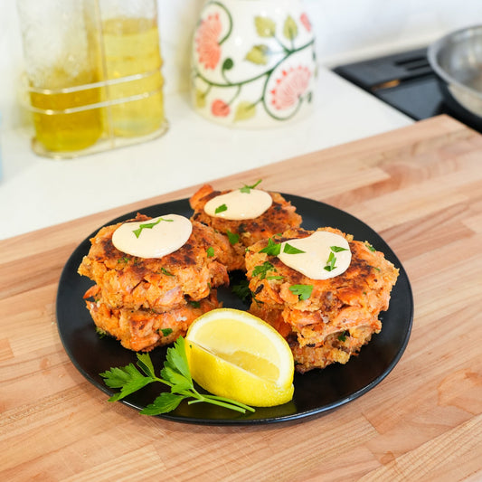 Close-up of salmon cakes showcasing sriracha aioli and parsley garnish with lemon wedge on black plate on wooden board