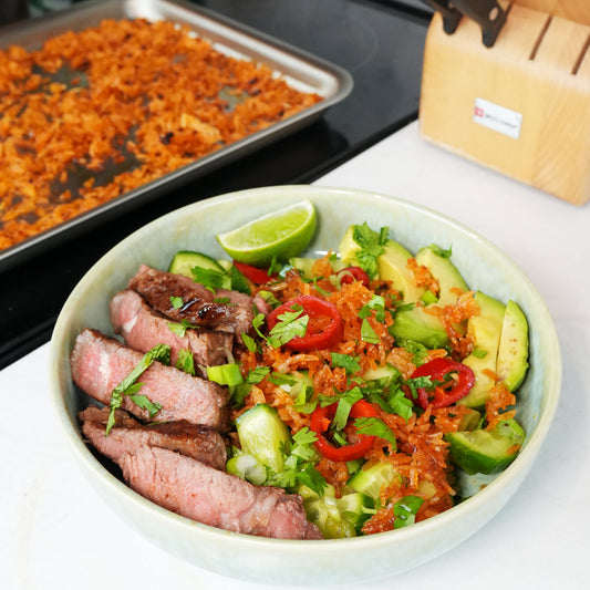 Bowl of sliced medium-rare steak over salad topped with crispy rice, avocado, red chiles and cilantro
