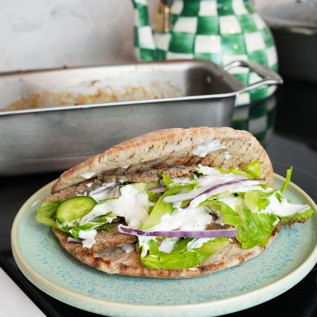 Oven-baked gyro in pita with dill yogurt sauce, lettuce, cucumber, and red onion on a plate; loaf pan in background