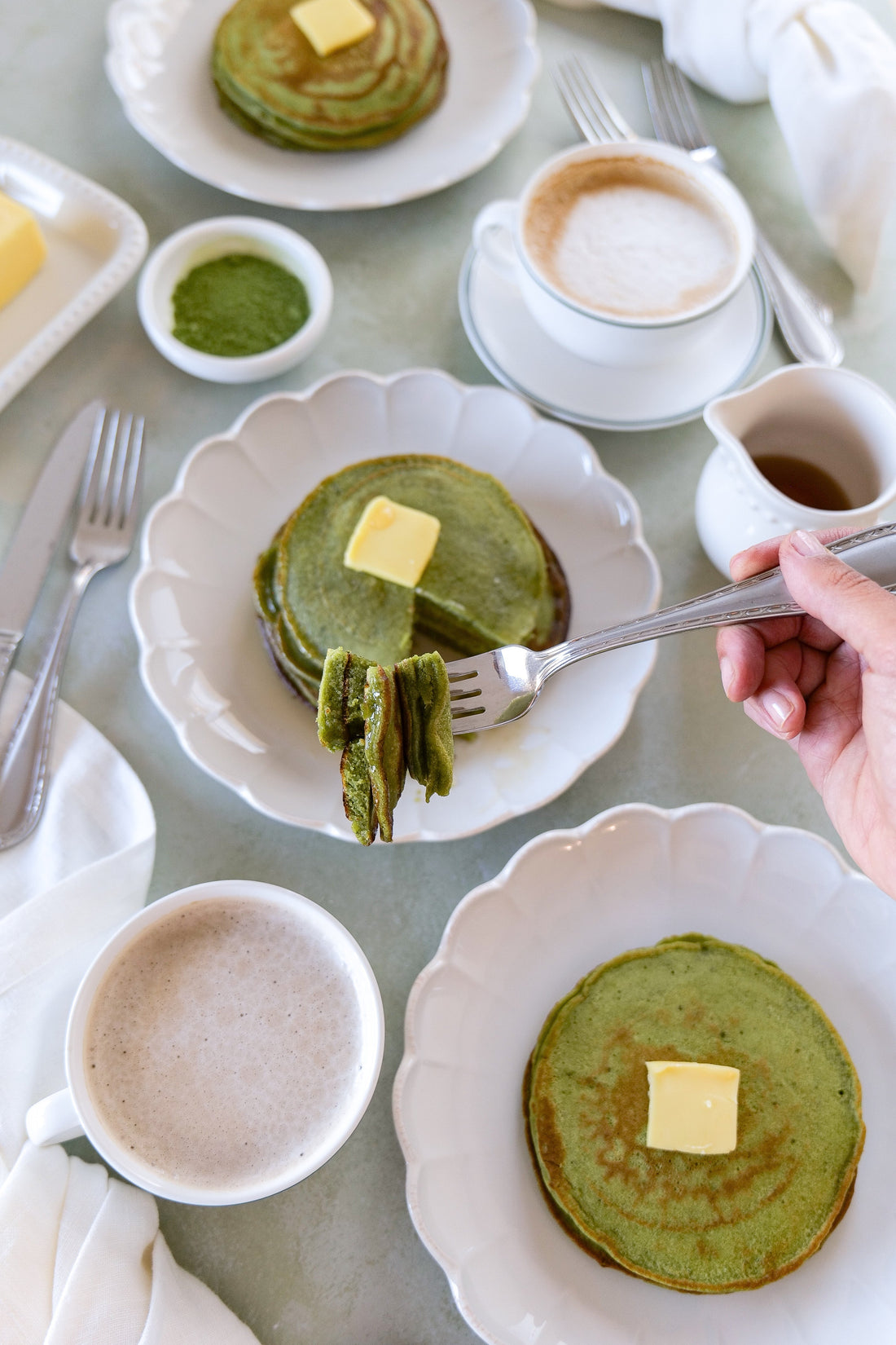 Matcha sourdough pancakes stacked with butter, a fork lifting a bite, served with coffee and a small pitcher of syrup on white plates