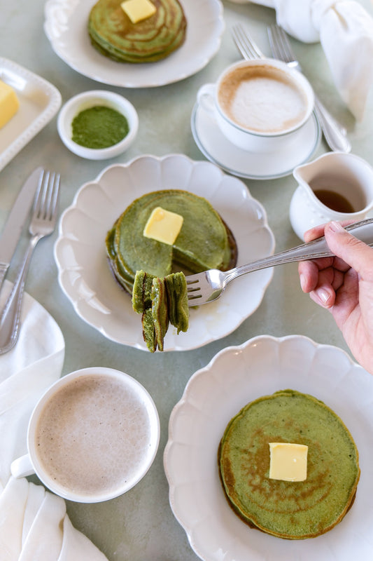 Matcha sourdough pancakes stacked with butter, a fork lifting a bite, served with coffee and a small pitcher of syrup on white plates