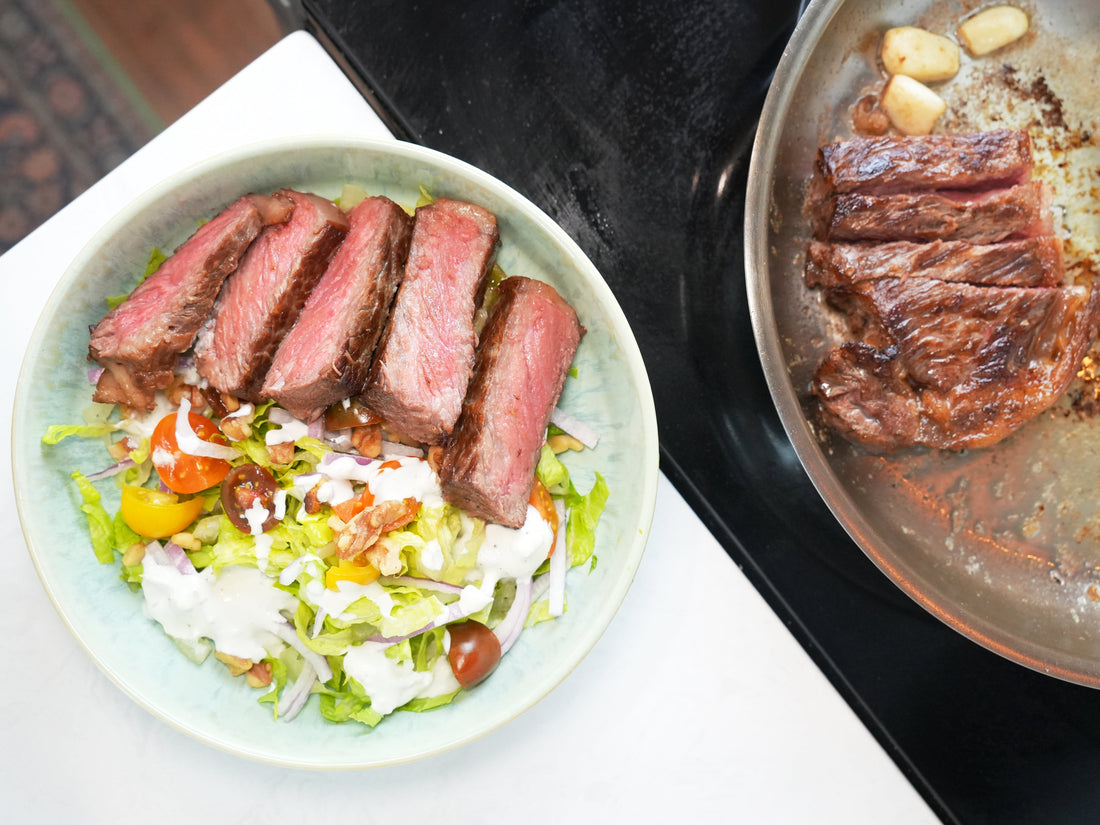 Sliced medium-rare steak on chopped salad with cherry tomatoes, walnuts and creamy dressing, pan of seared steak at right