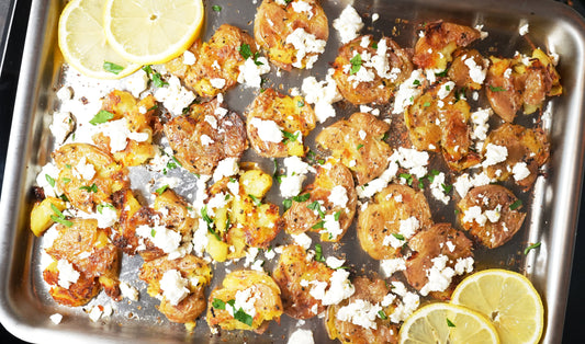 Crispy smashed baby potatoes topped with crumbled feta, chopped parsley, and lemon slices on a stainless steel roasting pan